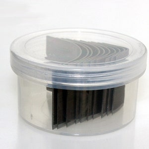 May include: A clear plastic container with a lid, containing a stack of thin, black, rectangular metal blades.