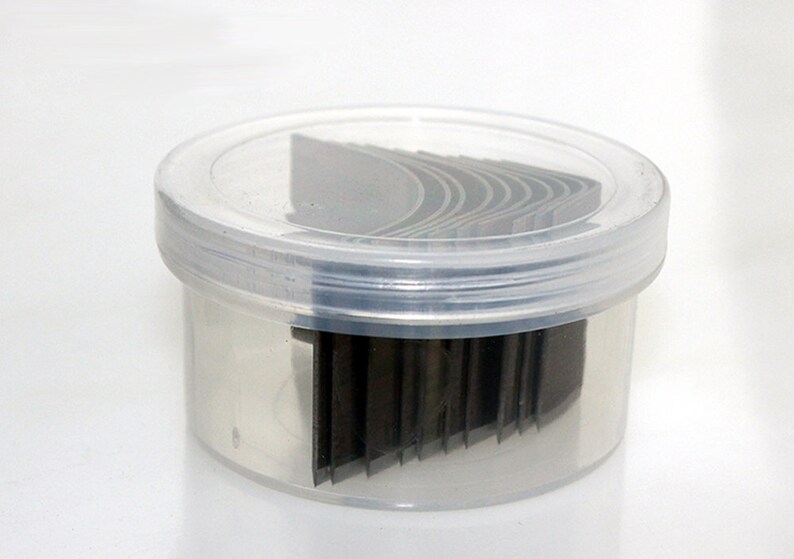 May include: A clear plastic container with a lid, containing a stack of thin, black, rectangular metal blades.