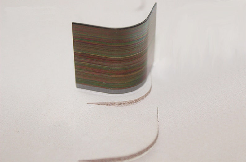 May include: A close-up of a curved, metallic ruler with a rainbow-like pattern of colored lines. The ruler is resting on a white surface, leaving a faint, curved line in the surface.