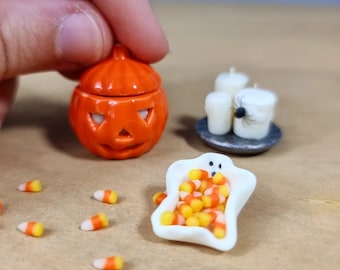 Può includere: Una piccola zucca di ceramica arancione con un coperchio, una ciotola fantasma di ceramica bianca piena di mais dolce e due candelieri di ceramica bianca con un ragno nero in cima.