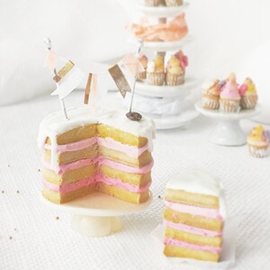 Puede incluir: Un pastel en miniatura de capas rosa y amarillo con glaseado blanco y una pequeña pancarta de banderas. El pastel está sobre un soporte para pastel blanco y hay otros pasteles en miniatura en el fondo.