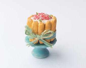 Peut inclure: Un gâteau miniature avec un glaçage blanc et des baies rouges sur le dessus, décoré d'un ruban vert et présenté sur un piédestal bleu.