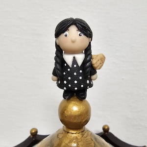 May include: A handcrafted figurine of a character with black braided hair, wearing a black dress with white polka dots and a white collar. The figure is holding a small object and stands atop a gold-colored sphere.