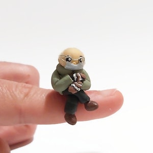 May include: A small, hand-crafted figurine of a person wearing a green jacket and glasses, sitting with their hands clasped in their lap.