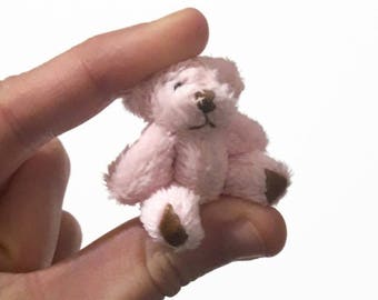 May include: A small pink teddy bear with brown eyes and a brown nose. The bear is sitting on a hand.