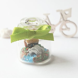 May include: A small glass jar with a green ribbon tied around the top. Inside the jar is a miniature scene of a house, a bridge, and a river.