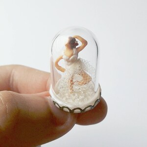 May include: A miniature ballerina figurine wearing a white tutu and a white veil is encased in a glass dome with a white and gold base.