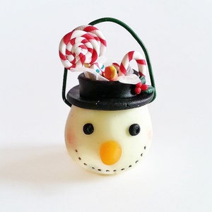 May include: A small snowman ornament with a black hat and a white face. The snowman is holding a candy cane and a lollipop.