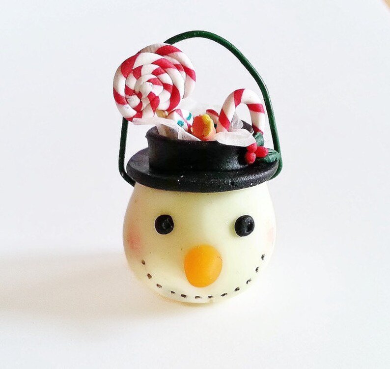May include: A small snowman ornament with a black hat and a white face. The snowman is holding a candy cane and a lollipop.