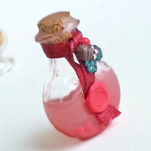 Puede incluir: Una peque&ntilde;a botella de vidrio llena de un l&iacute;quido rojo, coronada con un corcho y una cinta roja atada alrededor del cuello. La botella tiene un sello de cera roja y un peque&ntilde;o elemento decorativo verde y marr&oacute;n.