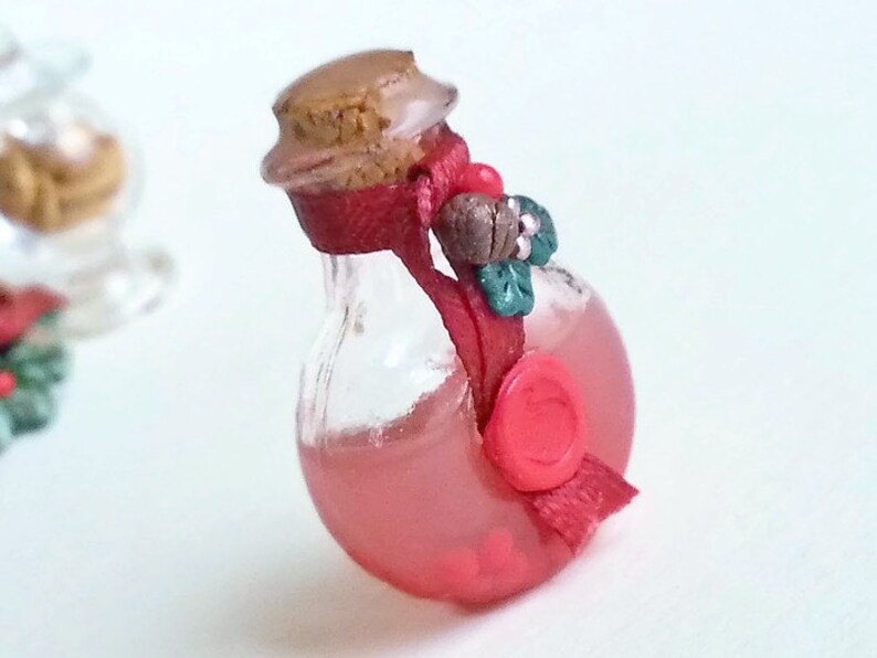 Puede incluir: Una peque&ntilde;a botella de vidrio llena de un l&iacute;quido rojo, coronada con un corcho y una cinta roja atada alrededor del cuello. La botella tiene un sello de cera roja y un peque&ntilde;o elemento decorativo verde y marr&oacute;n.