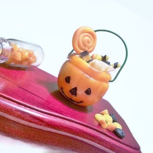 May include: A small orange pumpkin-shaped candy bucket with a green wire handle, filled with candy and a swirl lollipop. The bucket is sitting on a red wooden surface.