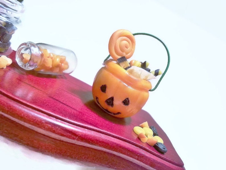 May include: A small orange pumpkin-shaped candy bucket with a green wire handle, filled with candy and a swirl lollipop. The bucket is sitting on a red wooden surface.