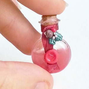 Puede incluir: Una peque&ntilde;a botella de vidrio con un tap&oacute;n de corcho, llena de un l&iacute;quido rosa. La botella est&aacute; decorada con una cinta roja, una rama de acebo verde y un sello de cera roja.