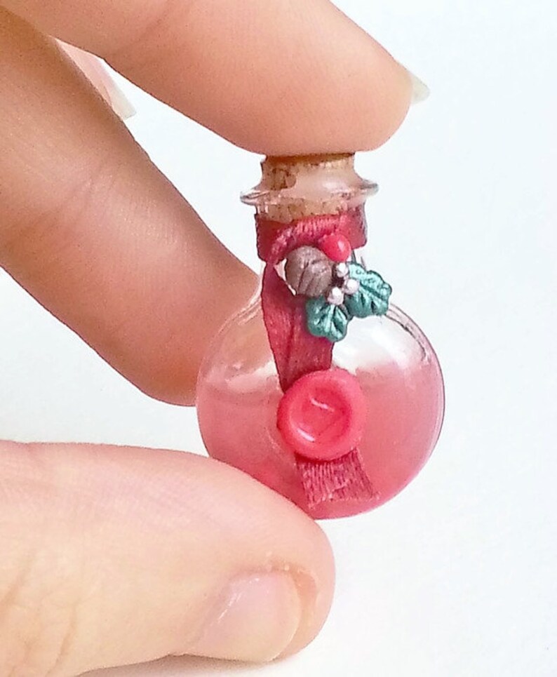 Puede incluir: Una peque&ntilde;a botella de vidrio con un tap&oacute;n de corcho, llena de un l&iacute;quido rosa. La botella est&aacute; decorada con una cinta roja, una rama de acebo verde y un sello de cera roja.