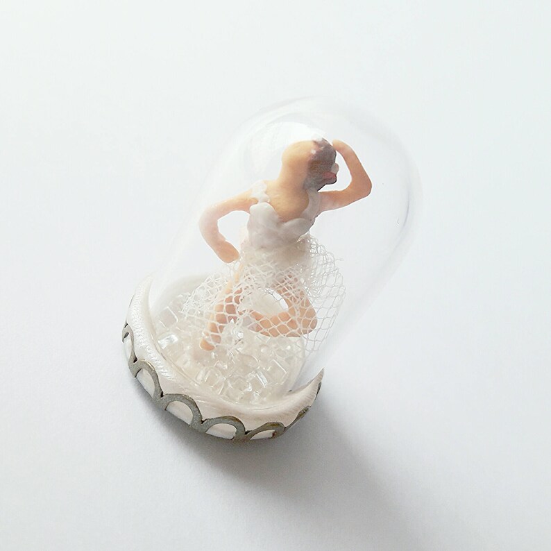 May include: A miniature ballerina figurine wearing a white tutu and a white veil is encased in a clear glass dome with a decorative base.