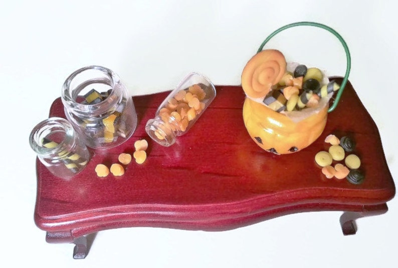 May include: A miniature wooden table with a pumpkin-shaped candy bucket, three glass jars filled with candy, and loose candy scattered on the table. The candy is a mix of orange and black.