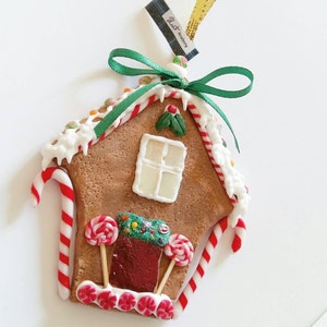 Puede incluir: Un adorno de casa de jengibre con glaseado blanco, bastones de caramelo rojo y blanco y decoraciones de caramelo de colores. La casa tiene un techo verde y una ventana blanca.