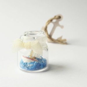 May include: A small glass jar with a white bow and a miniature sailboat inside. The jar is filled with blue and white material to represent the ocean.