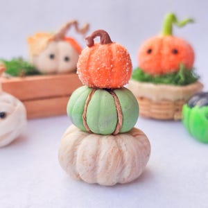 Può includere: Una pila di tre zucche decorative nei toni dell'arancione, del verde e del crema. La zucca superiore è arancione con una superficie strutturata, quella centrale è verde con dettagli in spago e quella inferiore è color crema. Altre zucche sono sullo sfondo.