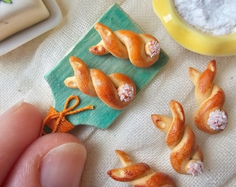 May include: Miniature baked goods, including twisted bread rolls with a light brown crust and white accents, are arranged on a green cutting board and a white surface. A small orange bow is visible. The scene evokes a bakery or food-related theme.