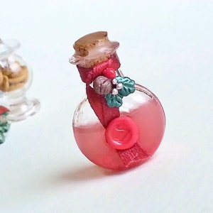 Puede incluir: Una peque&ntilde;a botella de vidrio con un l&iacute;quido rojo en su interior. La botella est&aacute; decorada con una cinta roja, una peque&ntilde;a pi&ntilde;a y hojas verdes. La botella tiene un tap&oacute;n de corcho y un sello de cera roja.