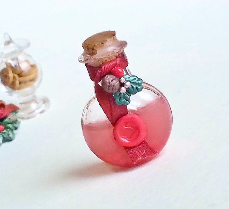 Puede incluir: Una peque&ntilde;a botella de vidrio con un l&iacute;quido rojo en su interior. La botella est&aacute; decorada con una cinta roja, una peque&ntilde;a pi&ntilde;a y hojas verdes. La botella tiene un tap&oacute;n de corcho y un sello de cera roja.