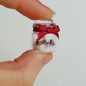 May include: A small glass jar with a red bow on top. Inside the jar is a miniature winter scene with a small house, trees, and snow.