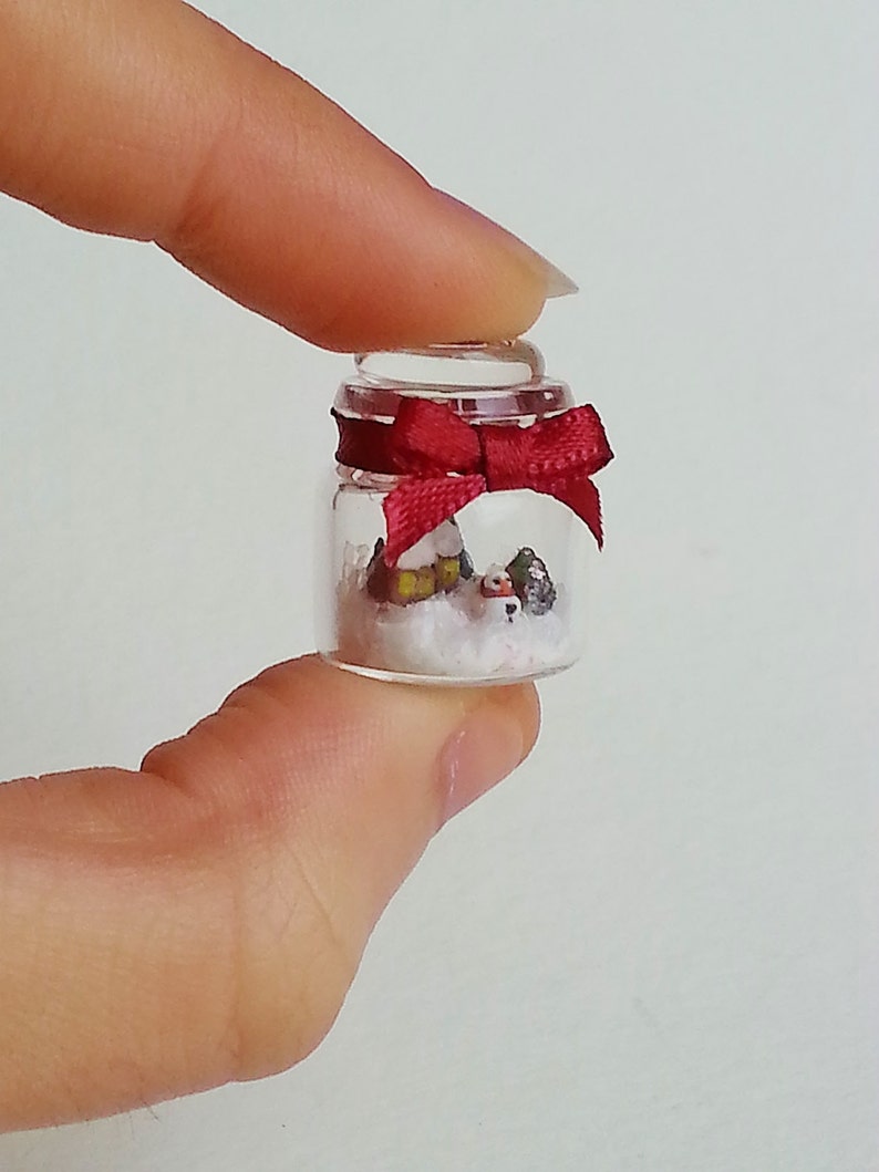 May include: A small glass jar with a red bow on top. Inside the jar is a miniature winter scene with a small house, trees, and snow.