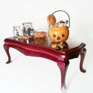 May include: A miniature wooden table with a red finish, decorated with a small orange pumpkin-shaped candy bucket, two glass jars filled with candy, and a scattering of candy on the table.