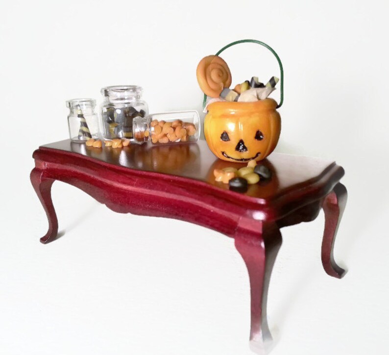 May include: A miniature wooden table with a red finish, decorated with a small orange pumpkin-shaped candy bucket, two glass jars filled with candy, and a scattering of candy on the table.