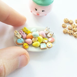 Puede incluir: Un plato blanco con una variedad de galletas y dulces en miniatura con temática de Pascua. Las galletas están decoradas con colores pastel y diseños de conejitos. Los dulces tienen forma de huevos y también están decorados con colores pastel.