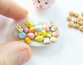 May include: A white plate with a variety of miniature Easter-themed cookies and candy. The cookies are decorated with pastel colors and bunny designs. The candy is in the shape of eggs and is also decorated with pastel colors.