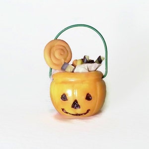 May include: A small orange pumpkin-shaped Halloween trick-or-treat bucket with a green wire handle. The bucket is filled with candy and a swirl lollipop.