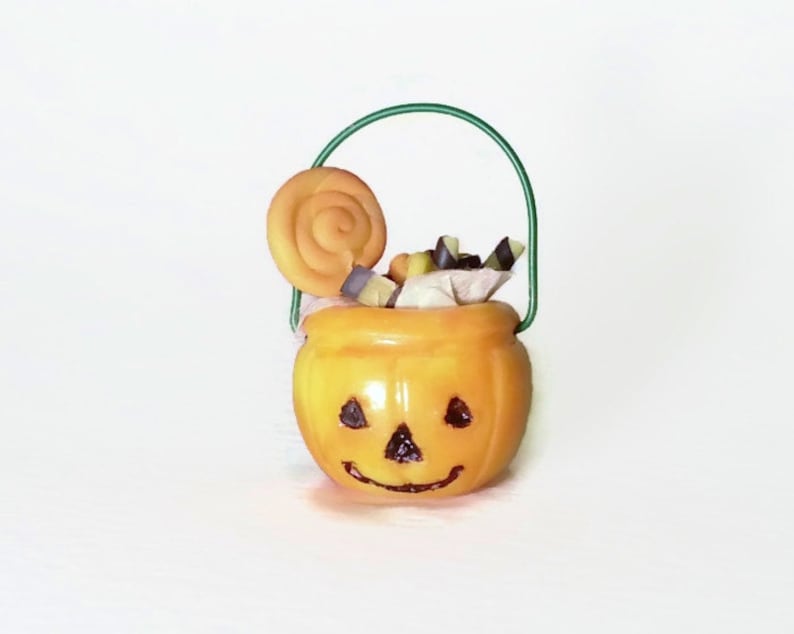 May include: A small orange pumpkin-shaped Halloween trick-or-treat bucket with a green wire handle. The bucket is filled with candy and a swirl lollipop.