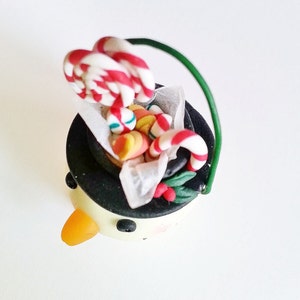 May include: A snowman ornament with a black top hat filled with candy canes and other colorful candies. The snowman has a yellow carrot nose and black eyes.