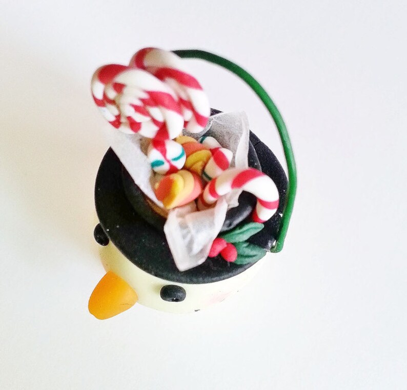 May include: A snowman ornament with a black top hat filled with candy canes and other colorful candies. The snowman has a yellow carrot nose and black eyes.