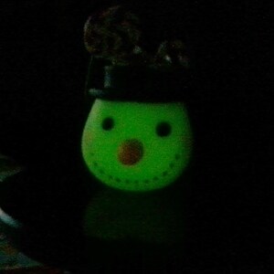 May include: A green snowman figurine with a black hat and a brown nose. The snowman is glowing in the dark.