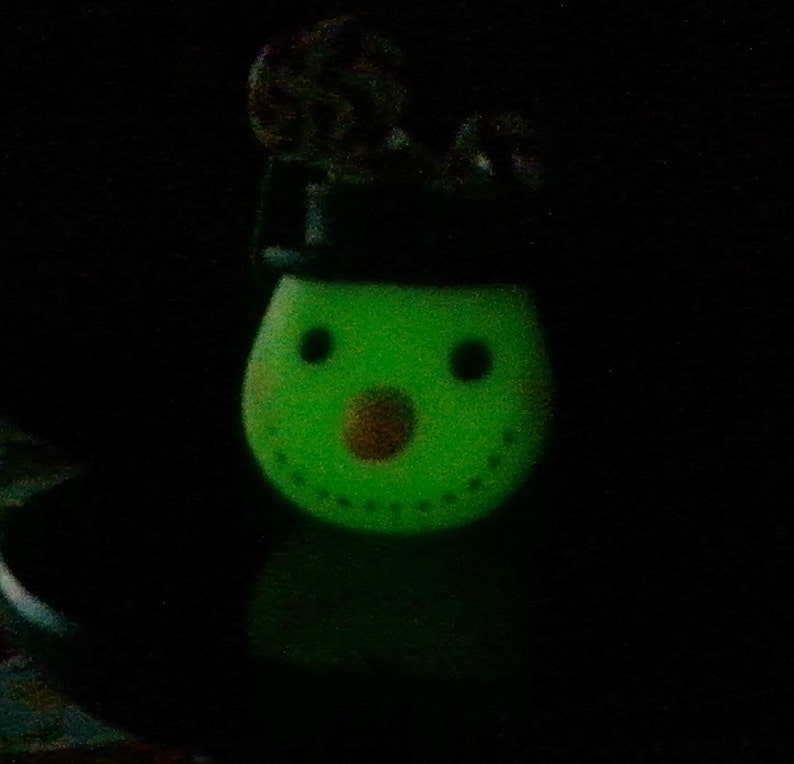 May include: A green snowman figurine with a black hat and a brown nose. The snowman is glowing in the dark.