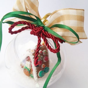 Puede incluir: Un adorno de vidrio transparente con una casa de jengibre en su interior. El adorno está decorado con una cinta roja, blanca y verde.