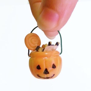 May include: A miniature orange pumpkin-shaped trick-or-treat bucket with a green wire handle. The bucket is filled with candy, including a swirl lollipop, candy corn, and black and white candy sticks.
