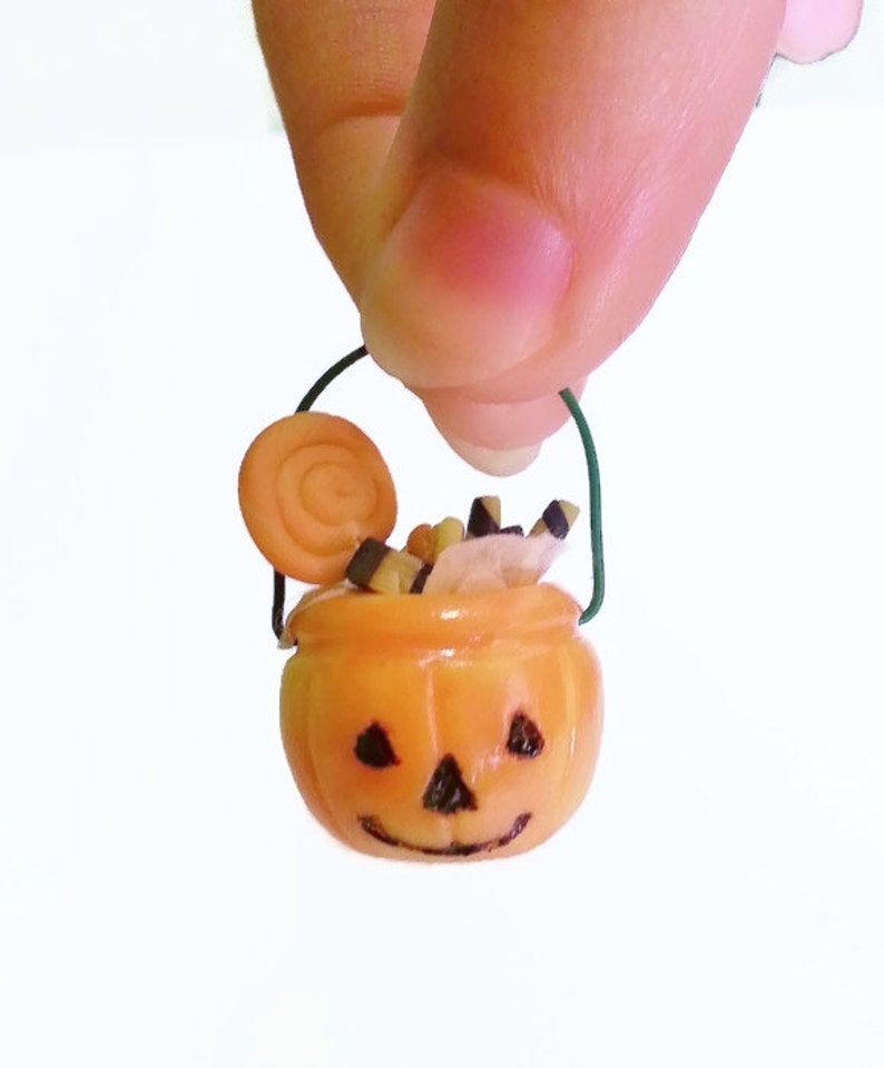 May include: A miniature orange pumpkin-shaped trick-or-treat bucket with a green wire handle. The bucket is filled with candy, including a swirl lollipop, candy corn, and black and white candy sticks.