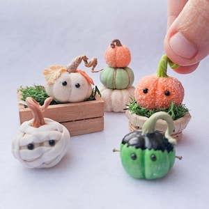 Puede incluir: Cinco calabazas en miniatura decoradas para Halloween. Una es una calabaza blanca con ojos saltones y envuelta en gasa blanca, otra es una calabaza verde con ojos saltones negros, una es una calabaza naranja con ojos saltones negros y un tallo marrón, y las dos últimas están apiladas una encima de la otra, una calabaza verde con un tallo marrón encima de una calabaza naranja con un tallo marrón.