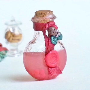 Puede incluir: Una peque&ntilde;a botella de vidrio con un tap&oacute;n de corcho y una cinta roja atada alrededor del cuello. La botella est&aacute; llena de un l&iacute;quido rosa y tiene un sello de cera roja en la parte delantera. La botella est&aacute; decorada con una rama de acebo.