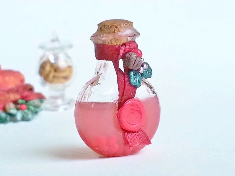 Puede incluir: Una peque&ntilde;a botella de vidrio con un tap&oacute;n de corcho y una cinta roja atada alrededor del cuello. La botella est&aacute; llena de un l&iacute;quido rosa y tiene un sello de cera roja en la parte delantera. La botella est&aacute; decorada con una rama de acebo.
