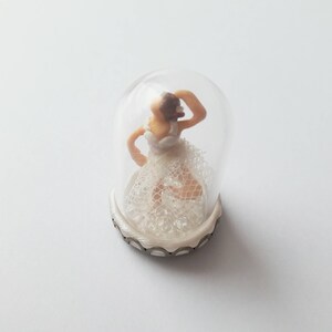 May include: A miniature ballerina figurine wearing a white lace dress is encased in a clear glass dome with a silver base.