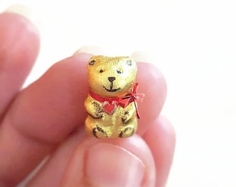 May include: A small gold teddy bear figurine with a red heart and a red bow. The bear is holding a red heart in its paws.