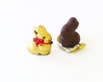 May include: Two chocolate bunny shaped candies wrapped in foil. One bunny is gold foil with a red ribbon and the other is silver foil with a brown chocolate bunny.