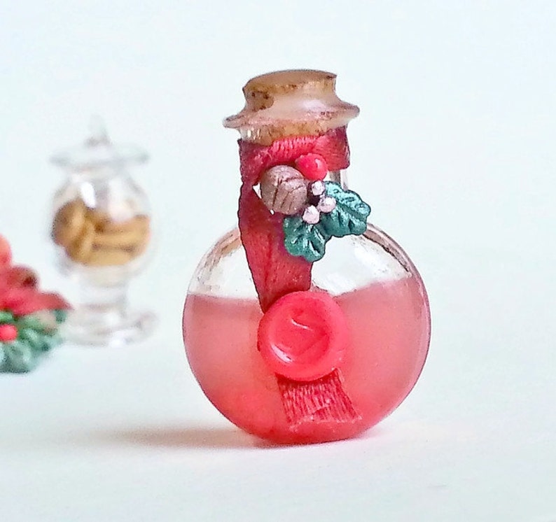 Puede incluir: Una peque&ntilde;a botella de vidrio con un l&iacute;quido rojo en su interior. La botella tiene un tap&oacute;n de corcho y est&aacute; decorada con una cinta roja y una rama de acebo verde.
