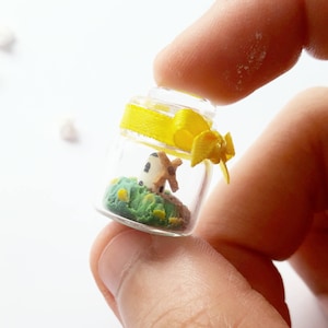 May include: A small glass jar with a yellow ribbon tied around the top. Inside the jar is a miniature windmill with green grass and yellow flowers.